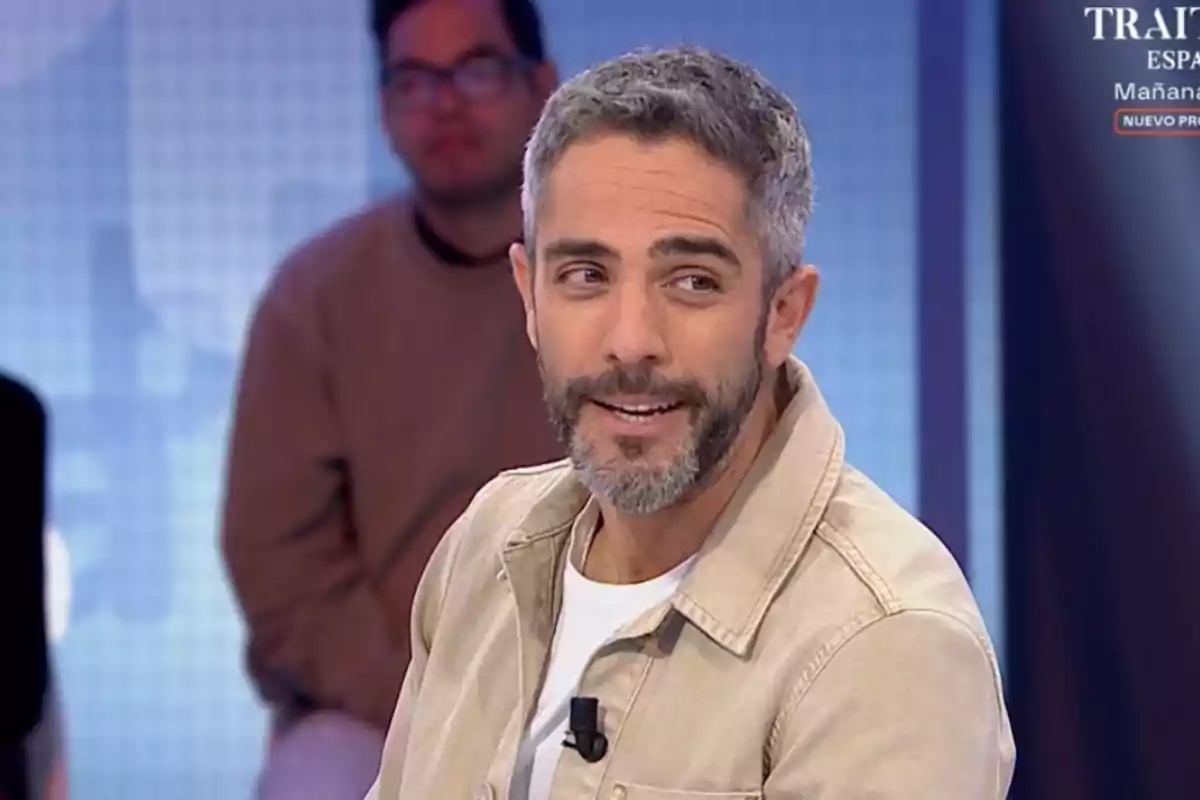Un home amb barba i cabell canós està somrient mentre participa en un programa de televisió amb un fons blau i una altra persona desenfocada darrere. Un home amb barba i cabell canós està somrient mentre participa en un programa de televisió amb un fons blau i una altra persona desenfocada darrere.
