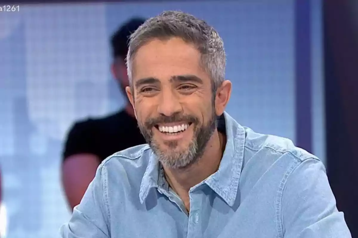 Un home somrient amb barba i camisa texana en un estudi de televisió. Un home somrient amb barba i camisa texana en un estudi de televisió.