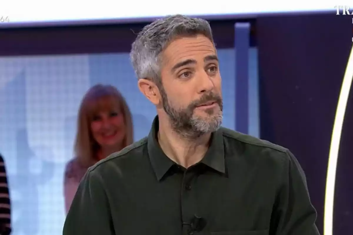 Un home amb barba i cabell canós és en un estudi de televisió, vestint una camisa fosca, mentre una dona somriu al fons. Un home amb barba i cabell canós és en un estudi de televisió, vestint una camisa fosca, mentre una dona somriu al fons.