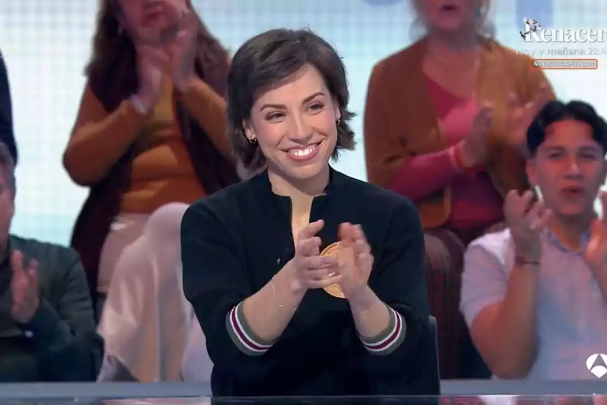 Una dona somrient aplaudeix mentre està asseguda en un programa de televisió amb persones aplaudint al fons. Una dona somrient aplaudeix mentre està asseguda en un programa de televisió amb persones aplaudint al fons.
