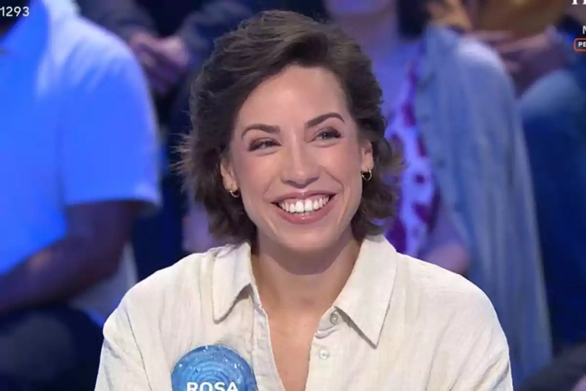 Dona jove de cabell curt i fosc somriu mentre participa en un programa de televisió, porta una camisa clara i una xapa blava amb el nom Rosa. Dona jove de cabell curt i fosc somriu mentre participa en un programa de televisió, porta una camisa clara i una xapa blava amb el nom Rosa.