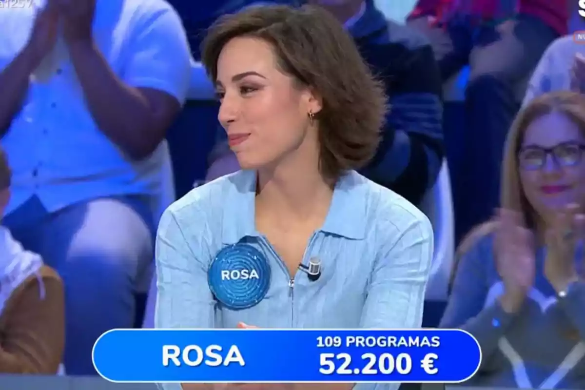 Una dona amb una camisa blava està asseguda en un programa de televisió, porta un distintiu amb el nom Una dona amb una camisa blava està asseguda en un programa de televisió, porta un distintiu amb el nom