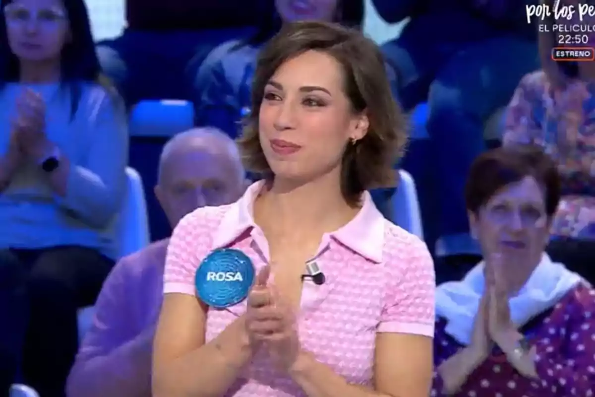 Una dona amb una samarreta rosa aplaudeix mentre està dreta en un estudi de televisió, envoltada de persones del públic. Una dona amb una samarreta rosa aplaudeix mentre està dreta en un estudi de televisió, envoltada de persones del públic.