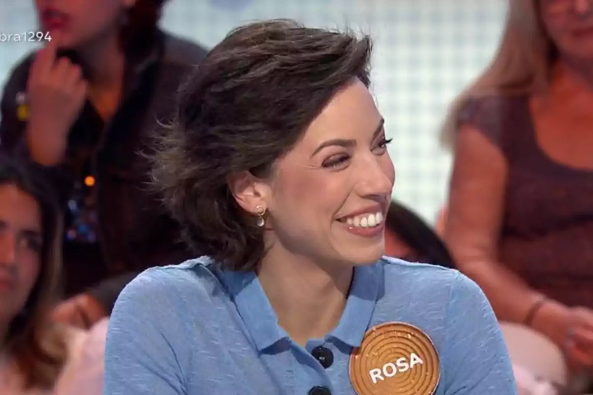 Dona somrient amb cabell curt i fosc porta una camisa blava i una insígnia amb el nom Rosa en un programa de televisió amb públic de fons Dona somrient amb cabell curt i fosc porta una camisa blava i una insígnia amb el nom Rosa en un programa de televisió amb públic de fons
