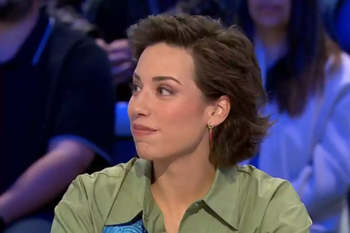 Una persona amb cabell curt i fosc, que porta una camisa verda i arracades, està asseguda en un entorn d'estudi de televisió, mirant cap a un costat. Una persona amb cabell curt i fosc, que porta una camisa verda i arracades, està asseguda en un entorn d'estudi de televisió, mirant cap a un costat.