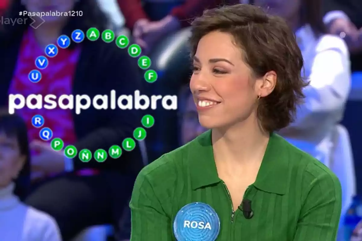 Una persona amb camisa verda i una etiqueta que diu "ROSA" està en un programa de televisió anomenat "Pasapalabra", amb un cercle de lletres al fons.