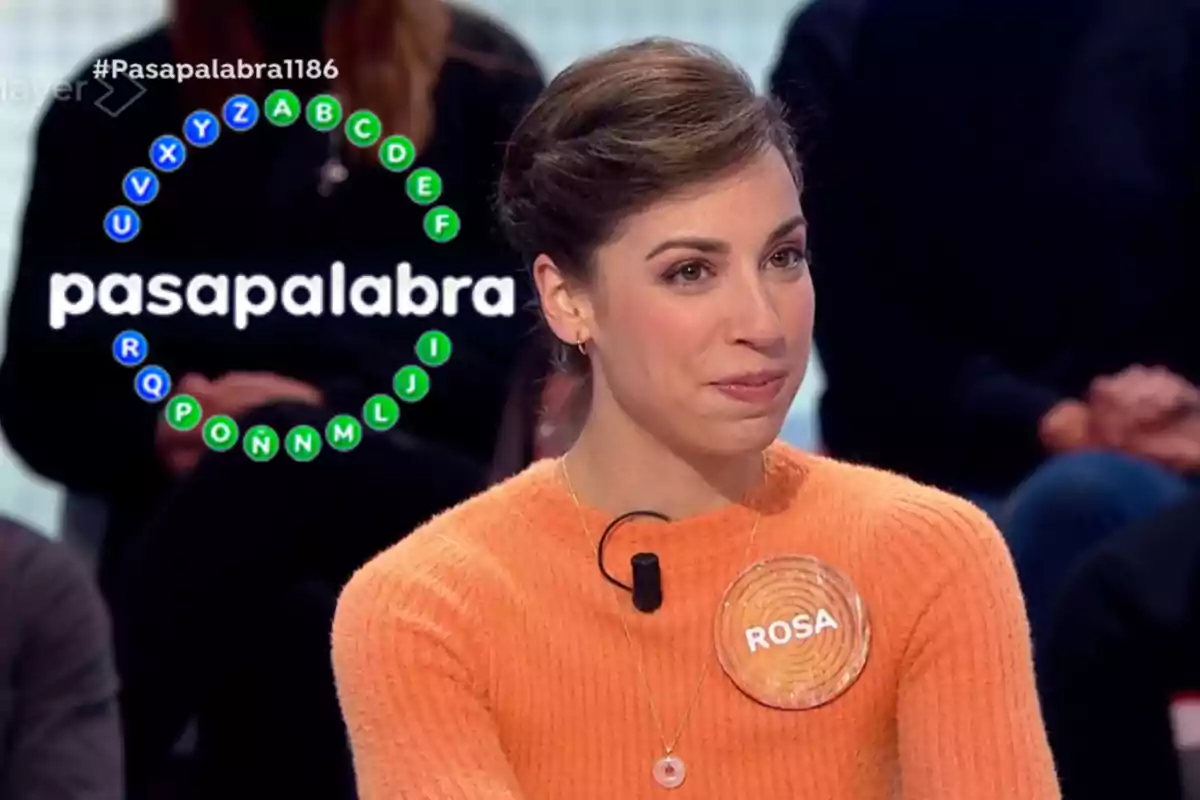 Una dona amb un suèter taronja participa en un programa de televisió anomenat "Pasapalabra" amb un cercle de lletres al fons.