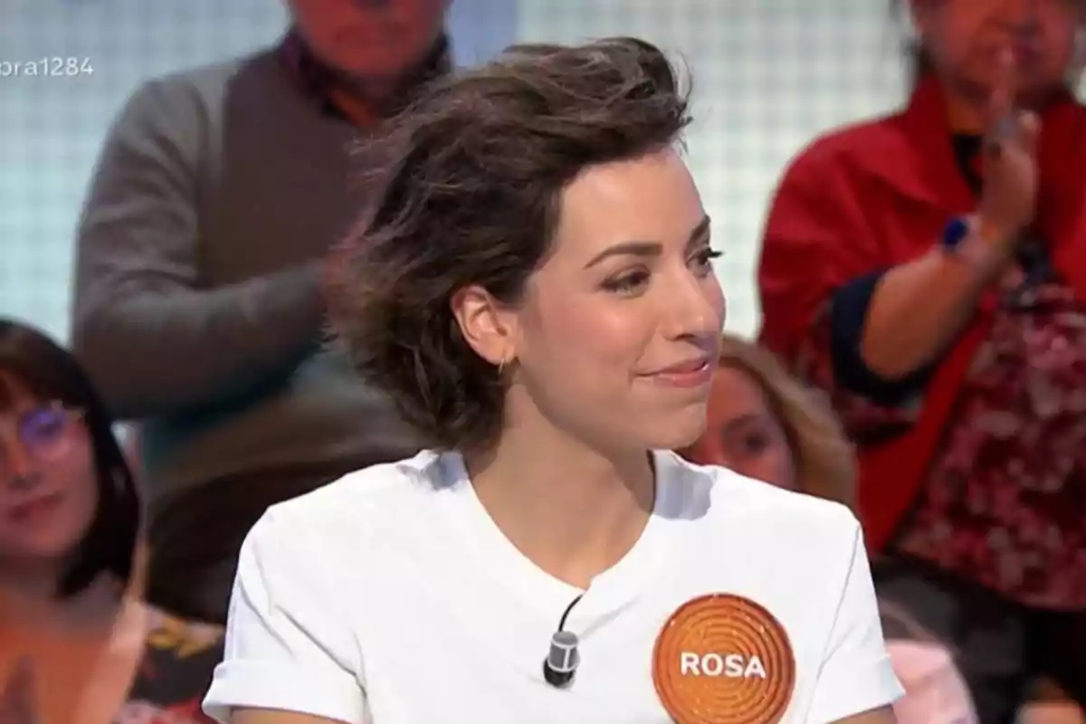 Dona jove amb cabell curt i fosc somriu mentre participa en un programa de televisió, porta una samarreta blanca amb un distintiu que diu Rosa i hi ha persones al fons aplaudint. Dona jove amb cabell curt i fosc somriu mentre participa en un programa de televisió, porta una samarreta blanca amb un distintiu que diu Rosa i hi ha persones al fons aplaudint.