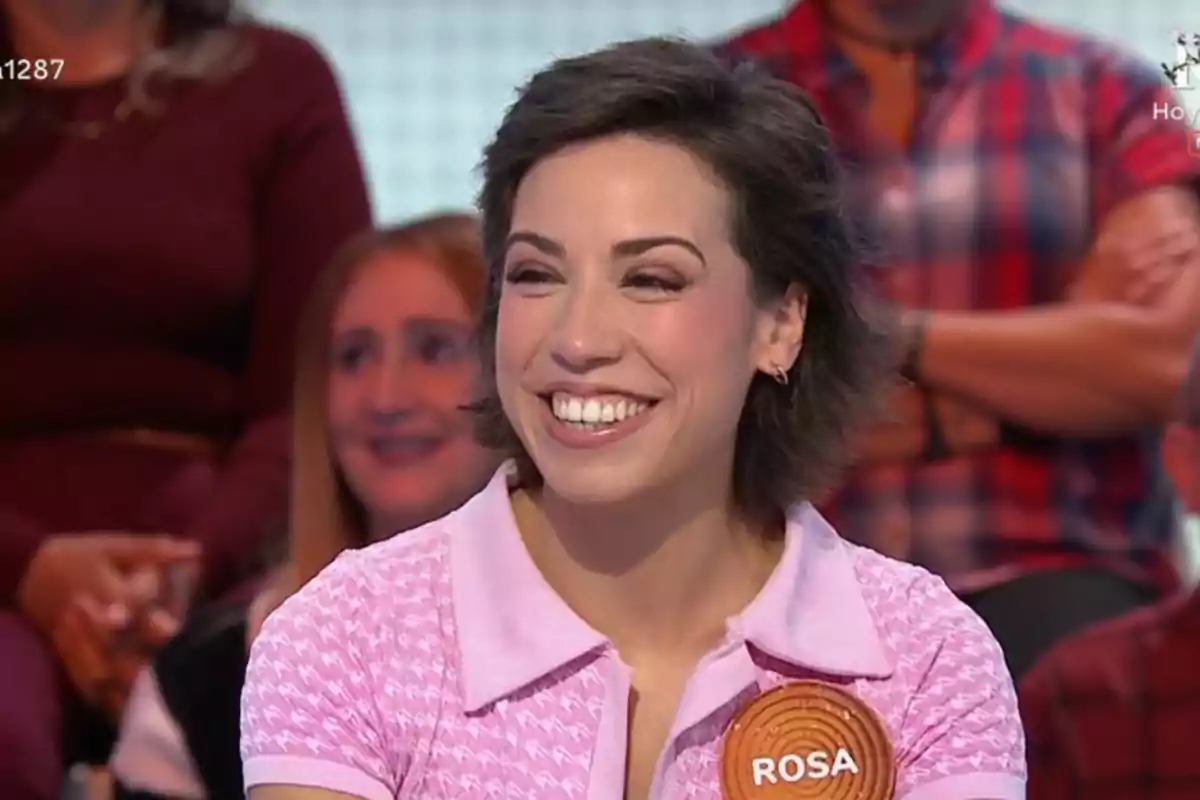 Dona somrient amb cabell curt i fosc porta una camisa rosa i una acreditació que diu Rosa mentre participa en un programa de televisió amb públic al fons. Dona somrient amb cabell curt i fosc porta una camisa rosa i una acreditació que diu Rosa mentre participa en un programa de televisió amb públic al fons.