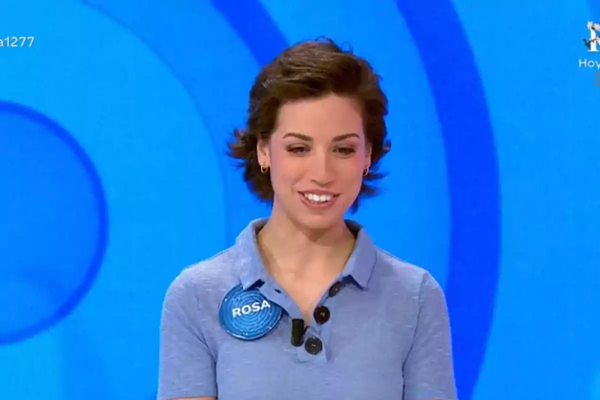 Dona jove amb cabell curt i fosc, somrient, vestida amb una camisa blau clar i un pin que diu Rosa, sobre un fons blau amb cercles concèntrics Dona jove amb cabell curt i fosc, somrient, vestida amb una camisa blau clar i un pin que diu Rosa, sobre un fons blau amb cercles concèntrics