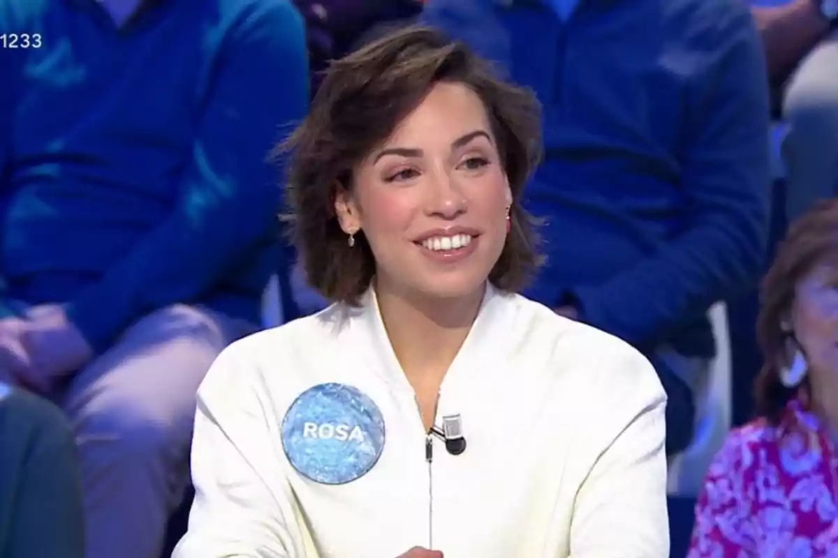 Una dona somrient amb cabell curt i fosc porta una jaqueta blanca i un micròfon en un programa de televisió amb públic al fons.