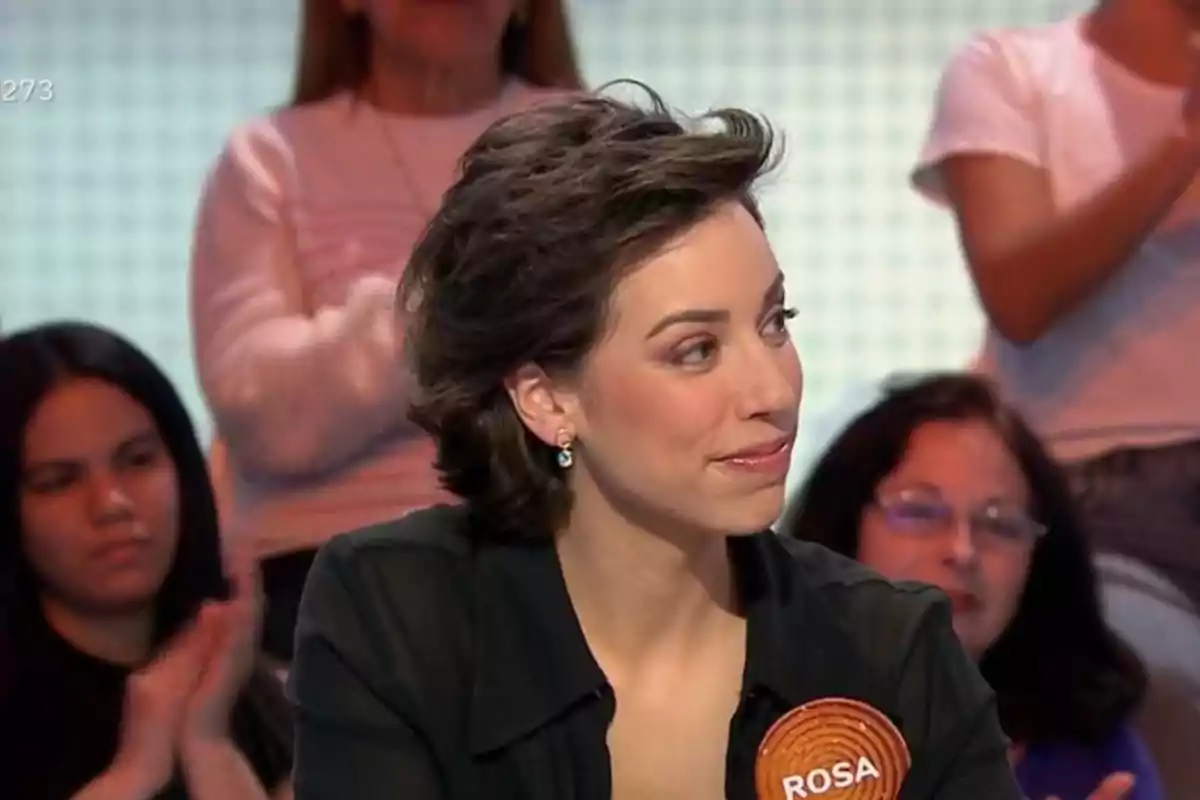 Una dona amb cabell curt i fosc, vestida amb una brusa negra, està asseguda en un programa de televisió amb un públic al fons. Una dona amb cabell curt i fosc, vestida amb una brusa negra, està asseguda en un programa de televisió amb un públic al fons.