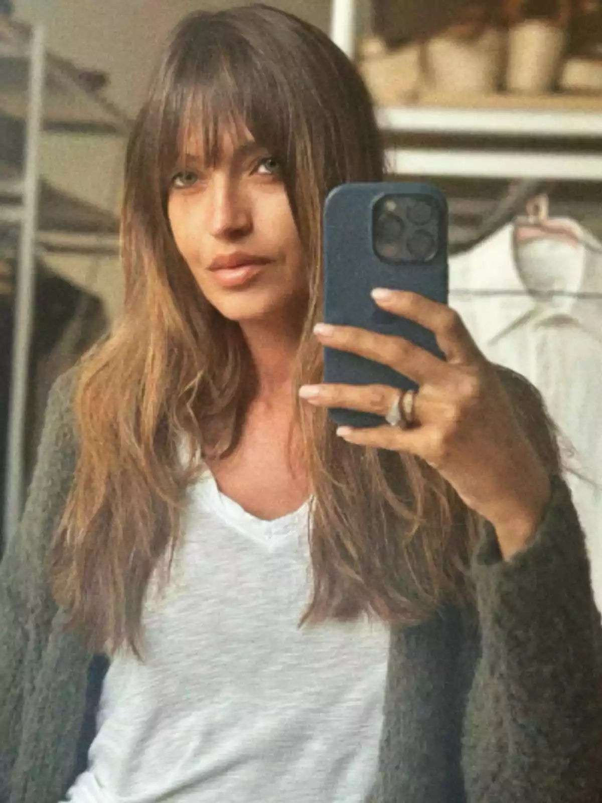 Sara Carbonero amb cabell llarg i serrell es pren una selfie davant d'un mirall, usant un telèfon mòbil amb funda blava, porta una samarreta blanca i un suèter verd, i al fons es veuen algunes peces penjades. Sara Carbonero amb cabell llarg i serrell es pren una selfie davant d'un mirall, usant un telèfon mòbil amb funda blava, porta una samarreta blanca i un suèter verd, i al fons es veuen algunes peces penjades.