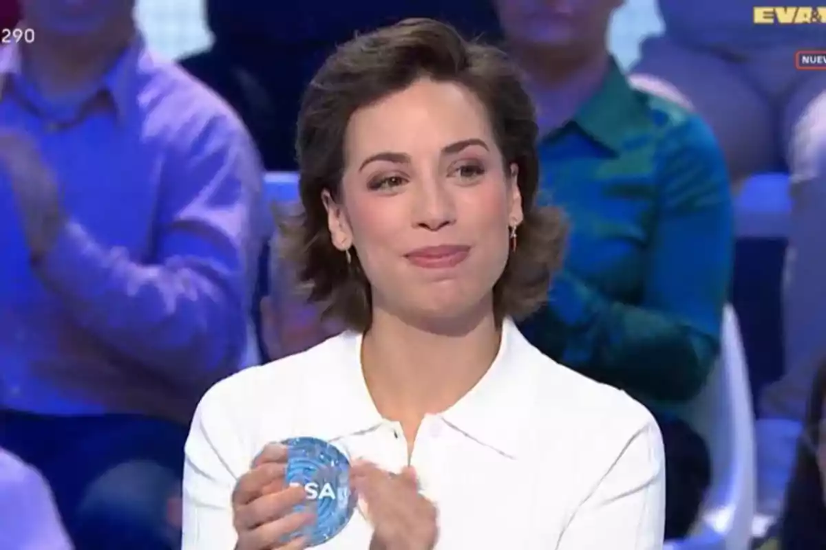 Dona amb cabell curt i camisa blanca somriu mentre sosté un objecte rodó blau en un programa de televisió amb públic de fons Dona amb cabell curt i camisa blanca somriu mentre sosté un objecte rodó blau en un programa de televisió amb públic de fons