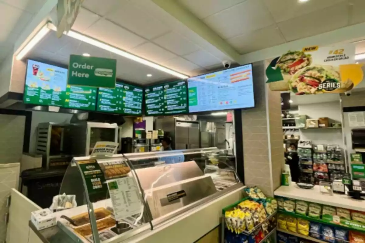 The image shows the interior of a fast-food restaurant - a Subway - with an order counter, digital menus at the top, and a variety of products on display, such as chips and drinks. The image shows the interior of a fast-food restaurant - a Subway - with an order counter, digital menus at the top, and a variety of products on display, such as chips and drinks.