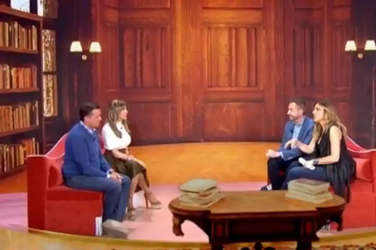 Un grup de persones està assegut en un plató de televisió amb una decoració que simula una biblioteca, amb prestatgeries plenes de llibres i una taula al centre. Un grup de persones està assegut en un plató de televisió amb una decoració que simula una biblioteca, amb prestatgeries plenes de llibres i una taula al centre.