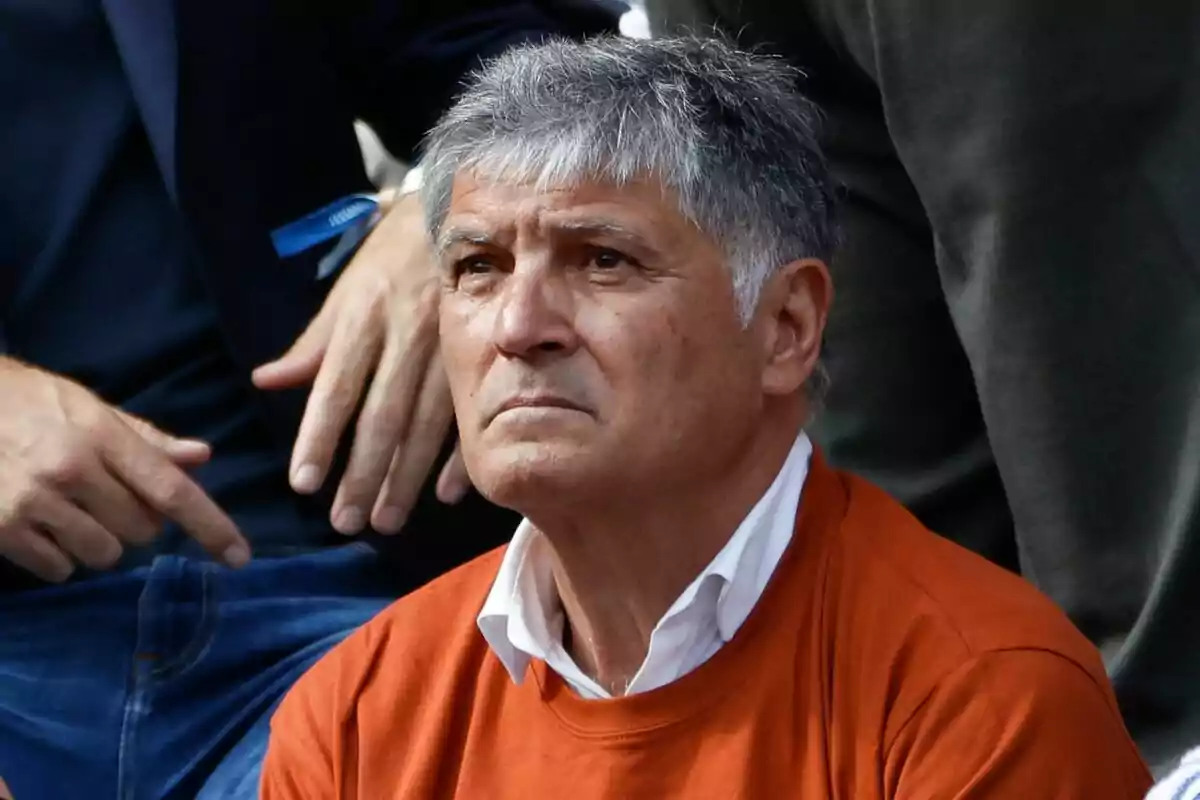 Gray-haired man with a serious expression wearing an orange sweater and a white jersey, surrounded by other people Gray-haired man with a serious expression wearing an orange sweater and a white jersey, surrounded by other people