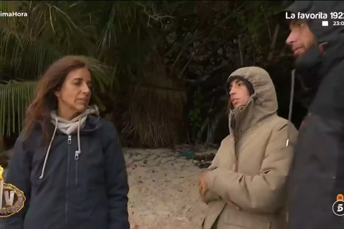 Tres persones abrigades conversen en una platja amb vegetació de fons. Tres persones abrigades conversen en una platja amb vegetació de fons.