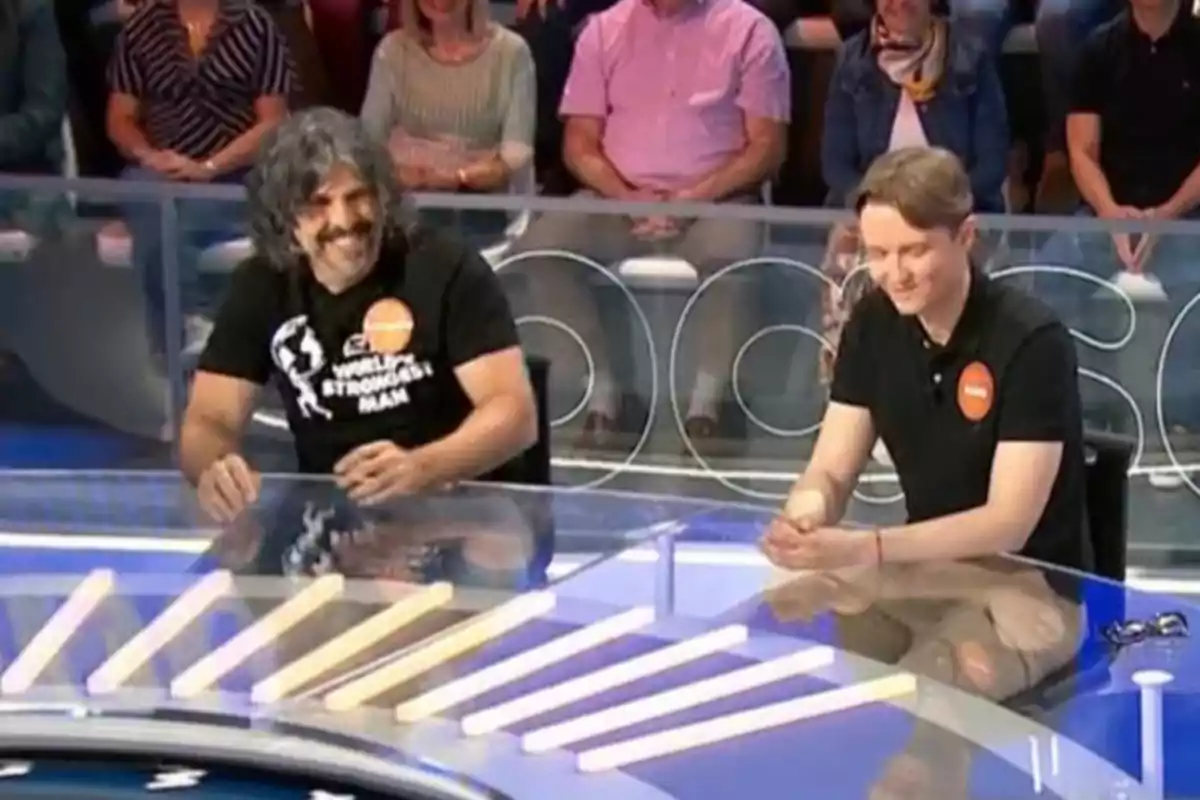Dos concursants asseguts en una taula transparent durant un programa de televisió amb públic al fons Dos concursants asseguts en una taula transparent durant un programa de televisió amb públic al fons