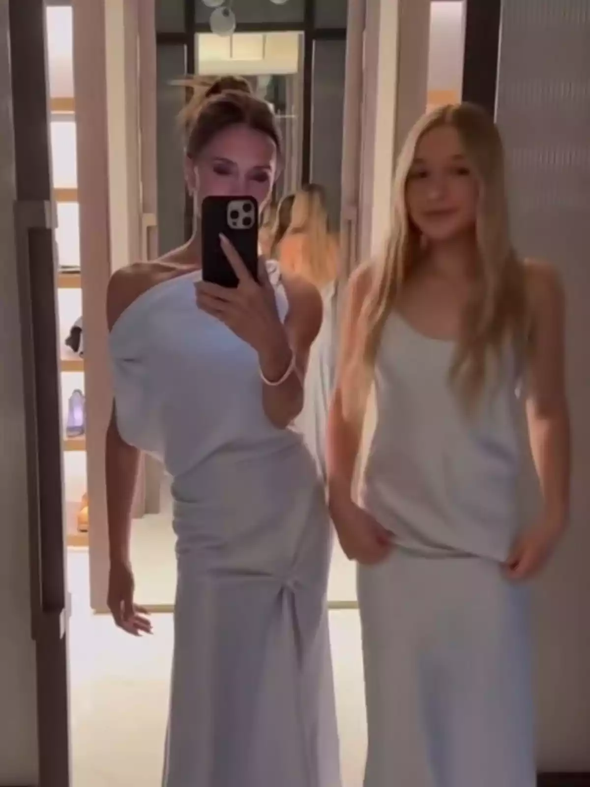 Dues persones posen davant d'un mirall, ambdues vestides amb elegants vestits blancs, mentre una d'elles sosté un telèfon mòbil per fer la foto. Dues persones posen davant d'un mirall, ambdues vestides amb elegants vestits blancs, mentre una d'elles sosté un telèfon mòbil per fer la foto.