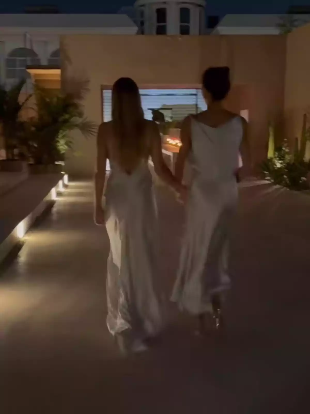 Dues persones amb vestits blancs caminen de la mà per un passadís il·luminat en un ambient exterior elegant. Dues persones amb vestits blancs caminen de la mà per un passadís il·luminat en un ambient exterior elegant.