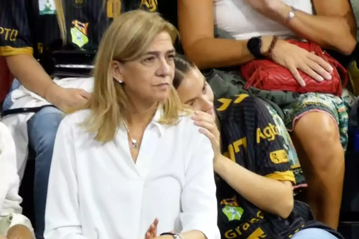 La infanta Cristina de cabell ros i camisa blanca està asseguda en un esdeveniment esportiu, amb persones al voltant vestint samarretes negres amb detalls grocs. La infanta Cristina de cabell ros i camisa blanca està asseguda en un esdeveniment esportiu, amb persones al voltant vestint samarretes negres amb detalls grocs.