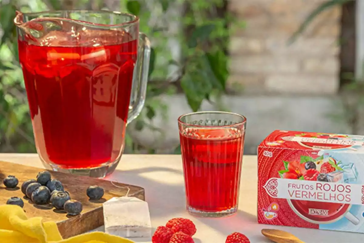 Una gerra i un got plens d'una beguda vermella són sobre una taula al costat d'una caixa de te de fruits vermells, nabius i gerds. Una gerra i un got plens d'una beguda vermella són sobre una taula al costat d'una caixa de te de fruits vermells, nabius i gerds.