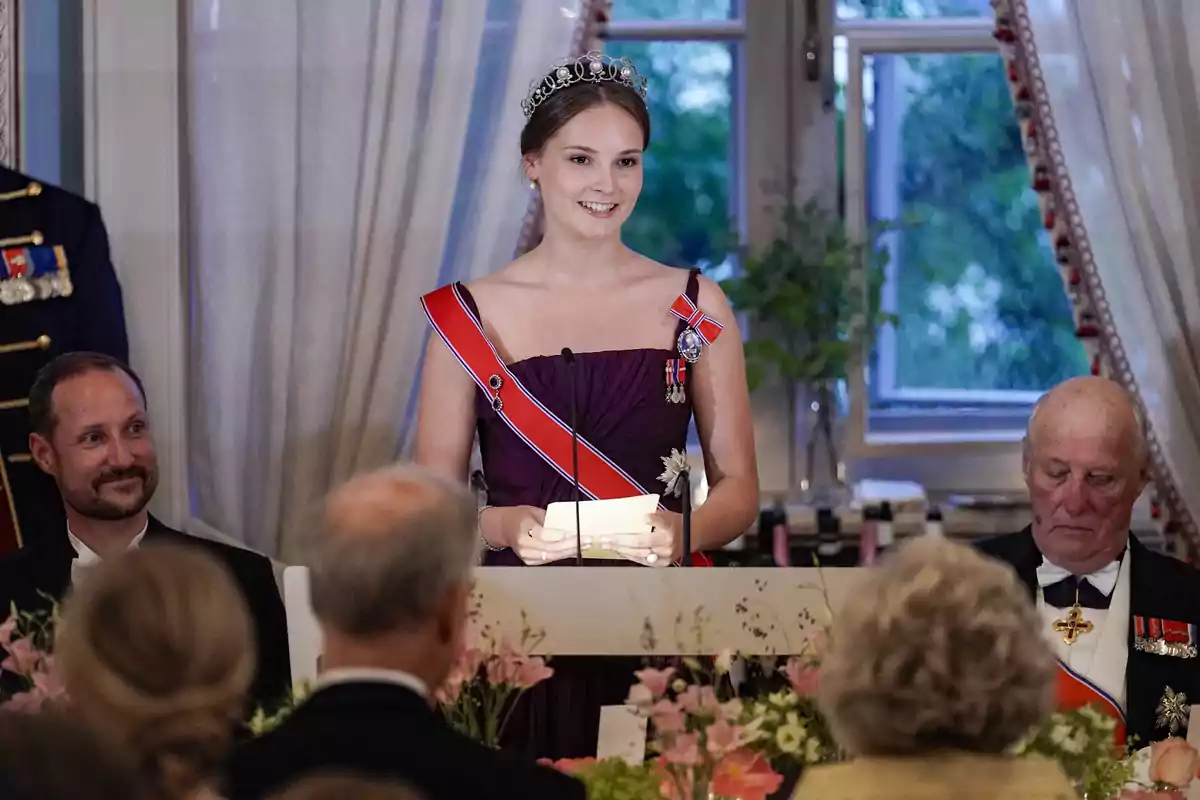 Ingrid Alexandra de Noruega amb una tiara i vestit formal fa un discurs en un esdeveniment elegant, envoltada de persones assegudes en una taula decorada amb flors. Ingrid Alexandra de Noruega amb una tiara i vestit formal fa un discurs en un esdeveniment elegant, envoltada de persones assegudes en una taula decorada amb flors.