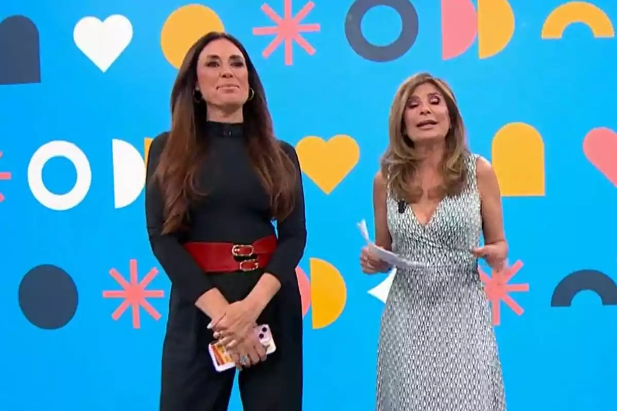 Isabel Rábago and Gema López standing on Espejo Público with a blue background decorated with geometric shapes and hearts, one wearing a black dress with a red belt and the other a black and white dress. Isabel Rábago and Gema López standing on Espejo Público with a blue background decorated with geometric shapes and hearts, one wearing a black dress with a red belt and the other a black and white dress.