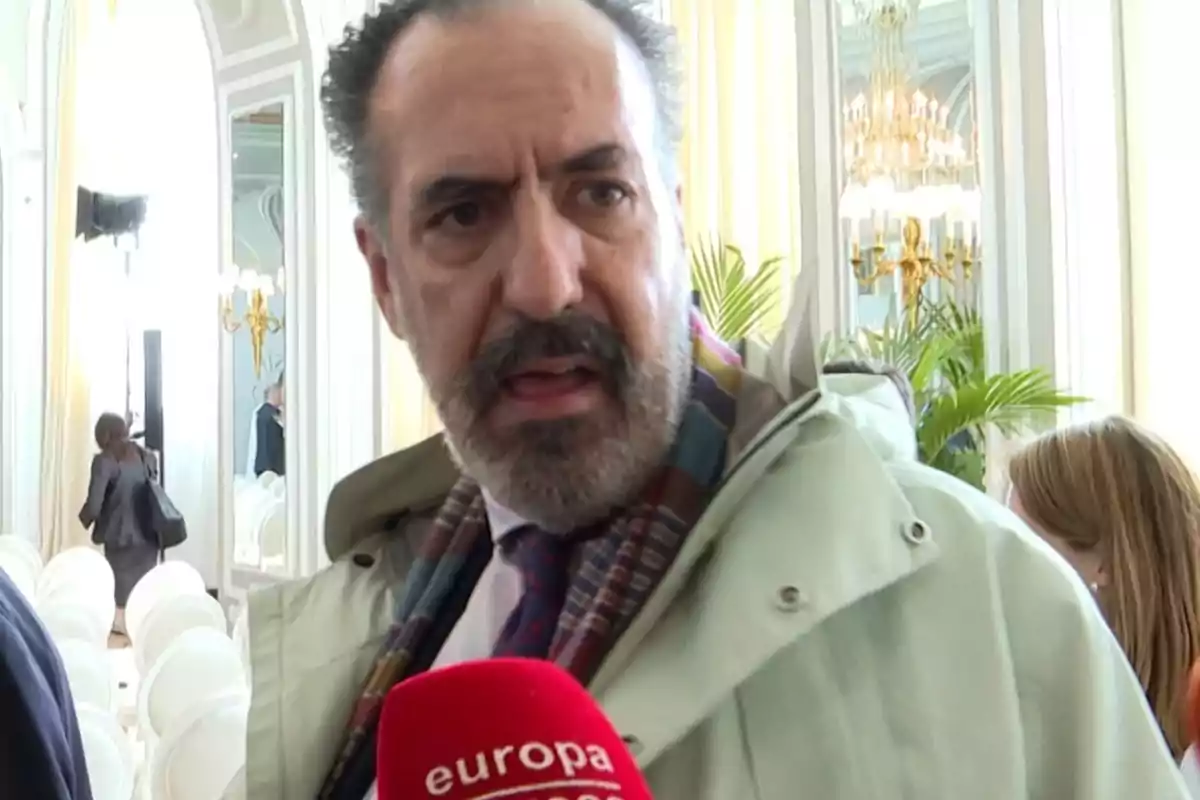 Un home amb barba i bigoti està parlant davant d'un micròfon vermell en una sala elegant amb miralls i canelobres. Un home amb barba i bigoti està parlant davant d'un micròfon vermell en una sala elegant amb miralls i canelobres.