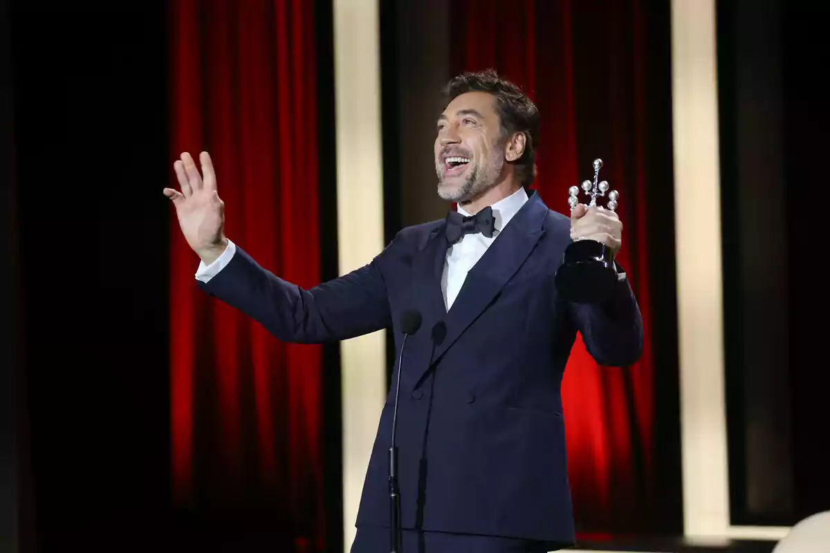 Javier Bardem on stage in a dark suit holding a trophy and smiling while speaking into the microphone. Javier Bardem on stage in a dark suit holding a trophy and smiling while speaking into the microphone.
