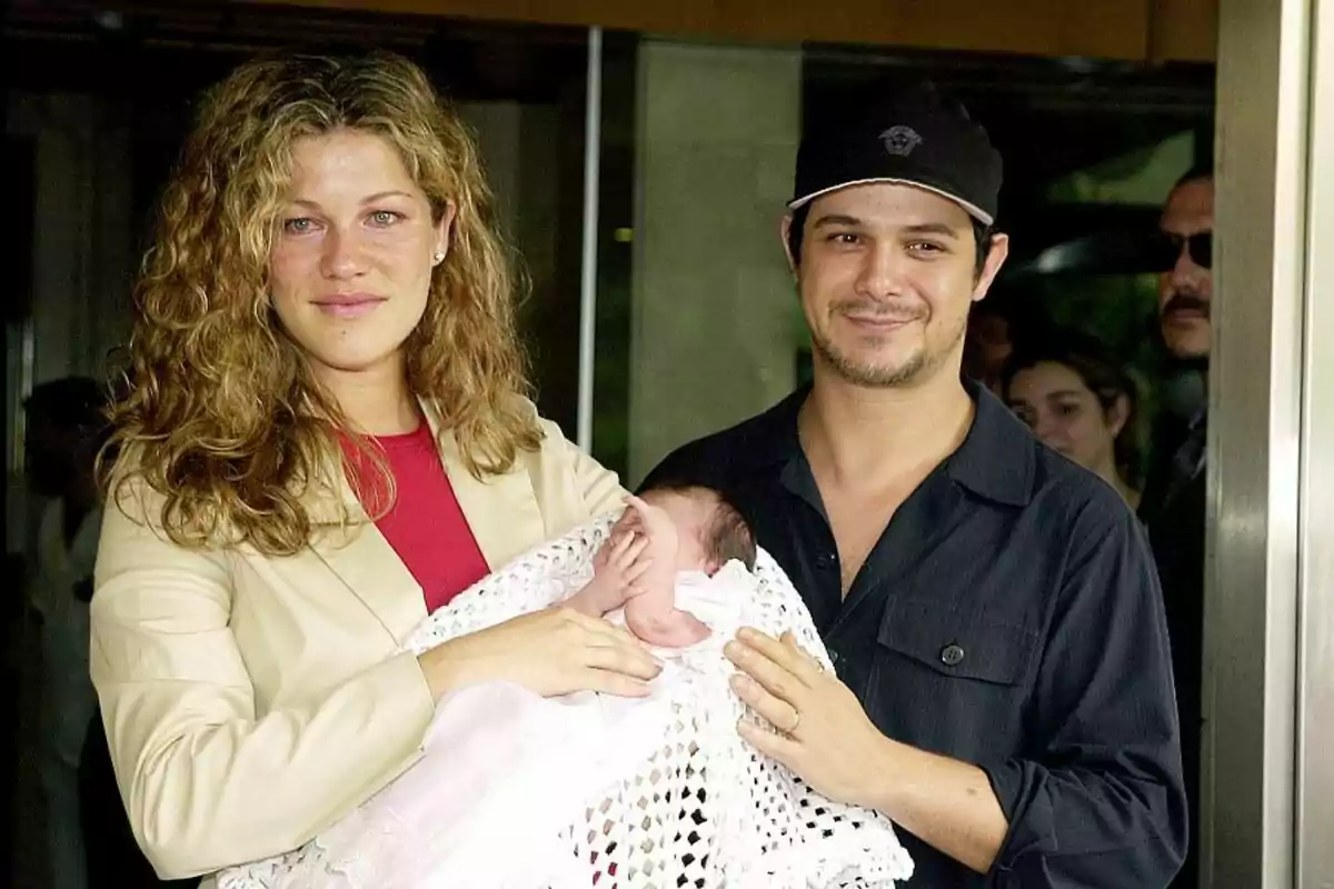 A smiling couple holds a baby wrapped in a white blanket as they pose for a photo in an outdoor setting. A smiling couple holds a baby wrapped in a white blanket as they pose for a photo in an outdoor setting.