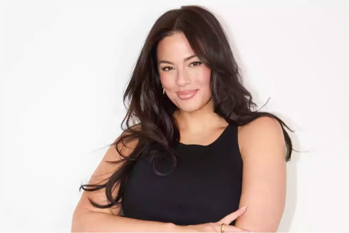 A person with long dark hair is wearing a black blouse and slightly smiling while crossing their arms in front of a white background. A person with long dark hair is wearing a black blouse and slightly smiling while crossing their arms in front of a white background.