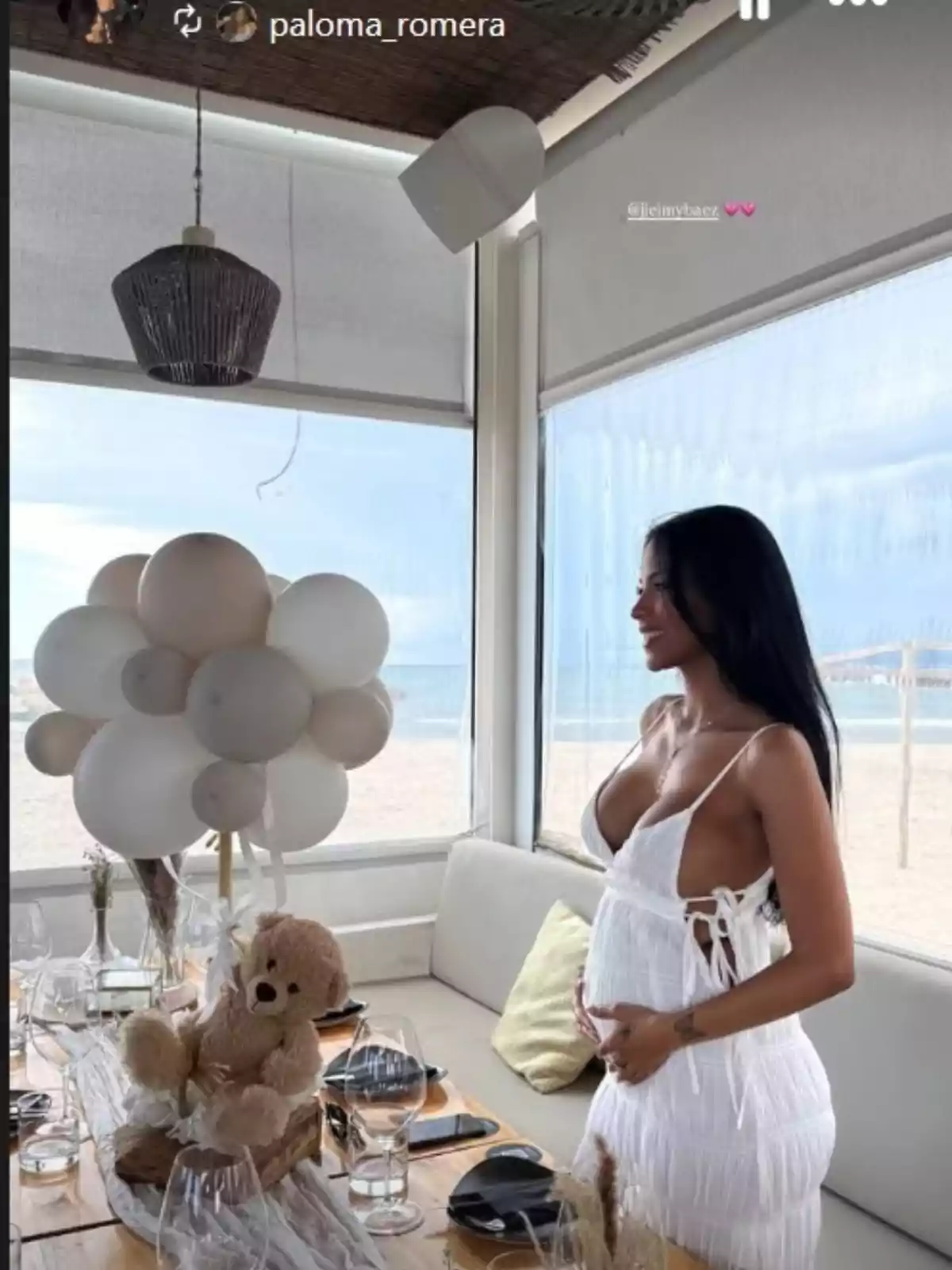 Jeimy Báez amb un vestit blanc està dempeus al costat d'una taula decorada amb globus blancs presumeix de panxa d'embarassada. Jeimy Báez amb un vestit blanc està dempeus al costat d'una taula decorada amb globus blancs presumeix de panxa d'embarassada.