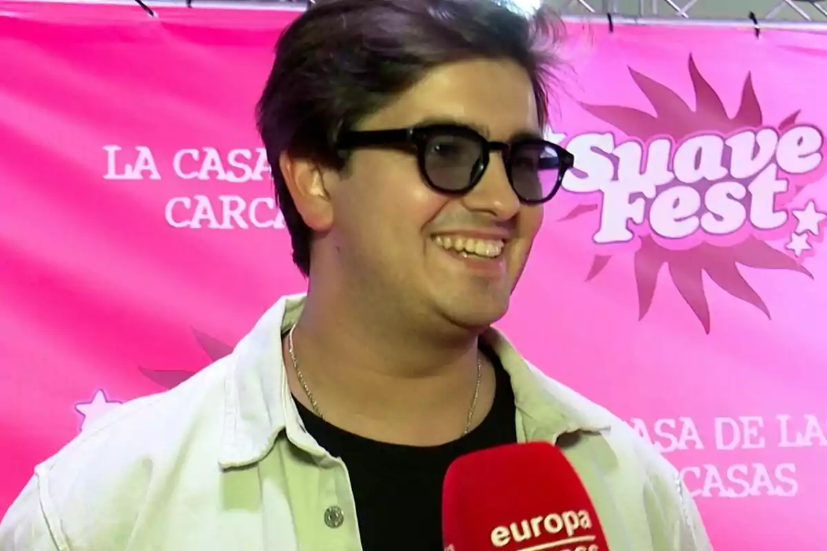 Smiling Jorge Bárcenas wearing sunglasses and a light jacket holds a red microphone in front of a pink background with the text 