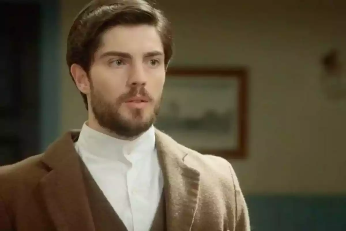 El actor Jorge Casat con barba y cabello oscuro, vestido con un abrigo marrón y una camisa blanca, mira hacia adelante con una expresión seria.