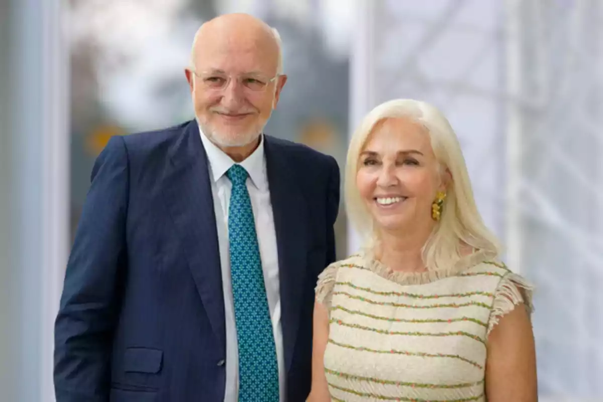 Elderly couple smiling as they pose for photo; man wearing blue suit with tie and woman in light dress with colorful details.