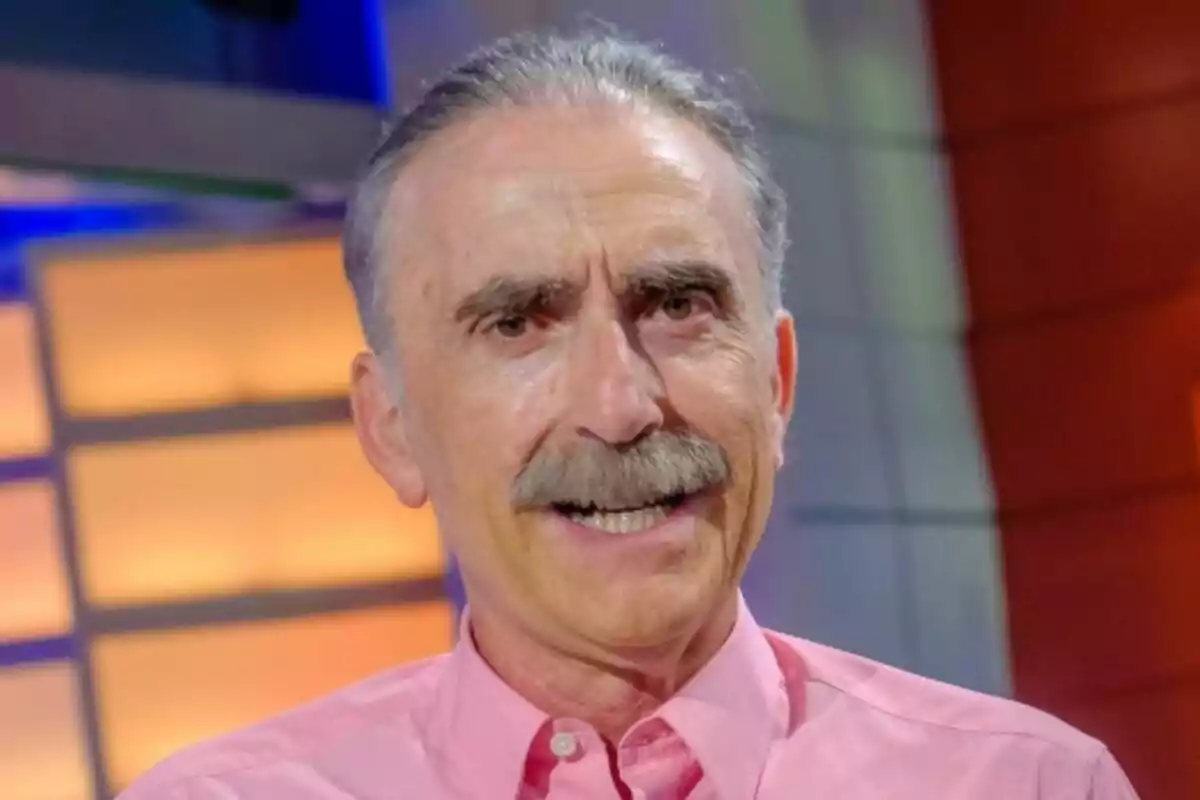 Juan y Medio de cabello canoso y bigote sonríe mientras lleva una camisa rosa en un entorno con luces cálidas en La tarde, aquí y ahora. Juan y Medio de cabello canoso y bigote sonríe mientras lleva una camisa rosa en un entorno con luces cálidas en La tarde, aquí y ahora.