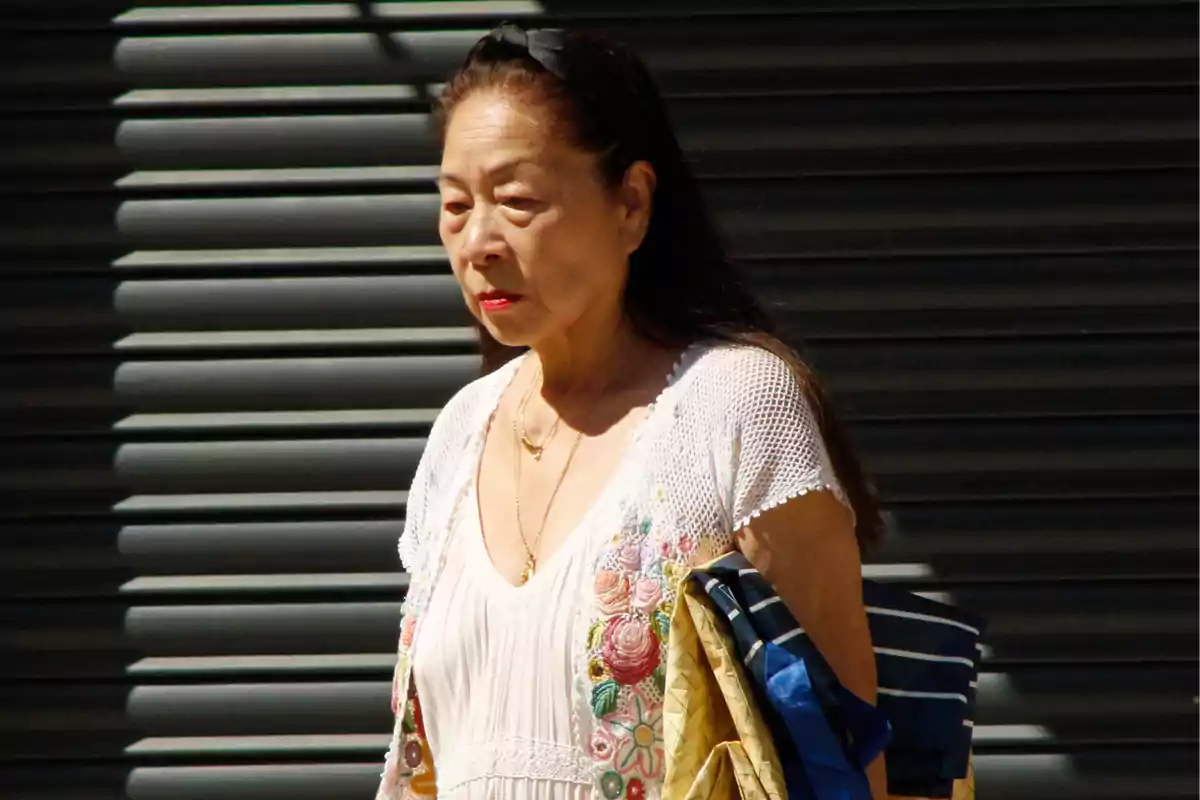 Junko caminant davant d'una persiana metàl·lica amb roba acolorida al braç.