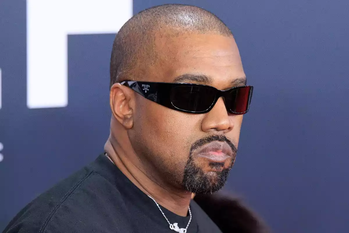 Kanye West wearing black sunglasses and a beard, dressed in a black t-jersey, at an event with a dark blue background. Kanye West wearing black sunglasses and a beard, dressed in a black t-jersey, at an event with a dark blue background.