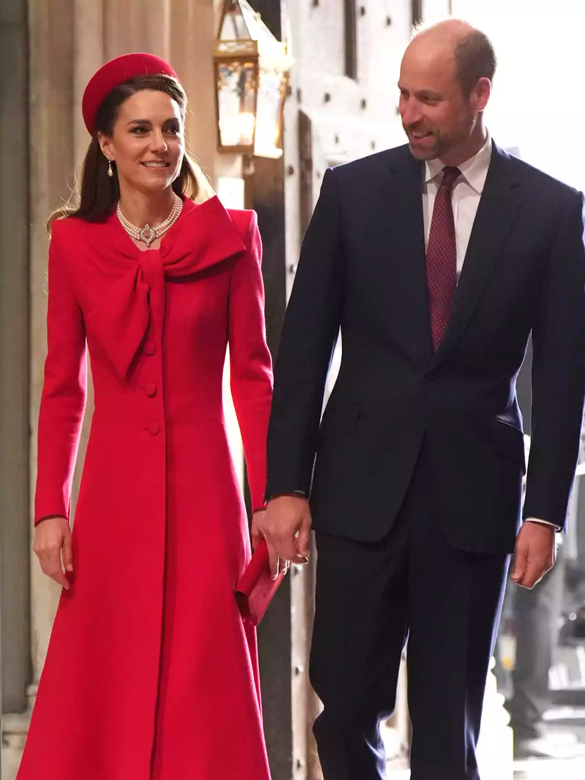 Kate Middleton i el príncep Guillem elegantment vestida camina de la mà en un esdeveniment formal, ella porta un vestit vermell amb un barret a joc i ell porta un vestit fosc amb corbata.