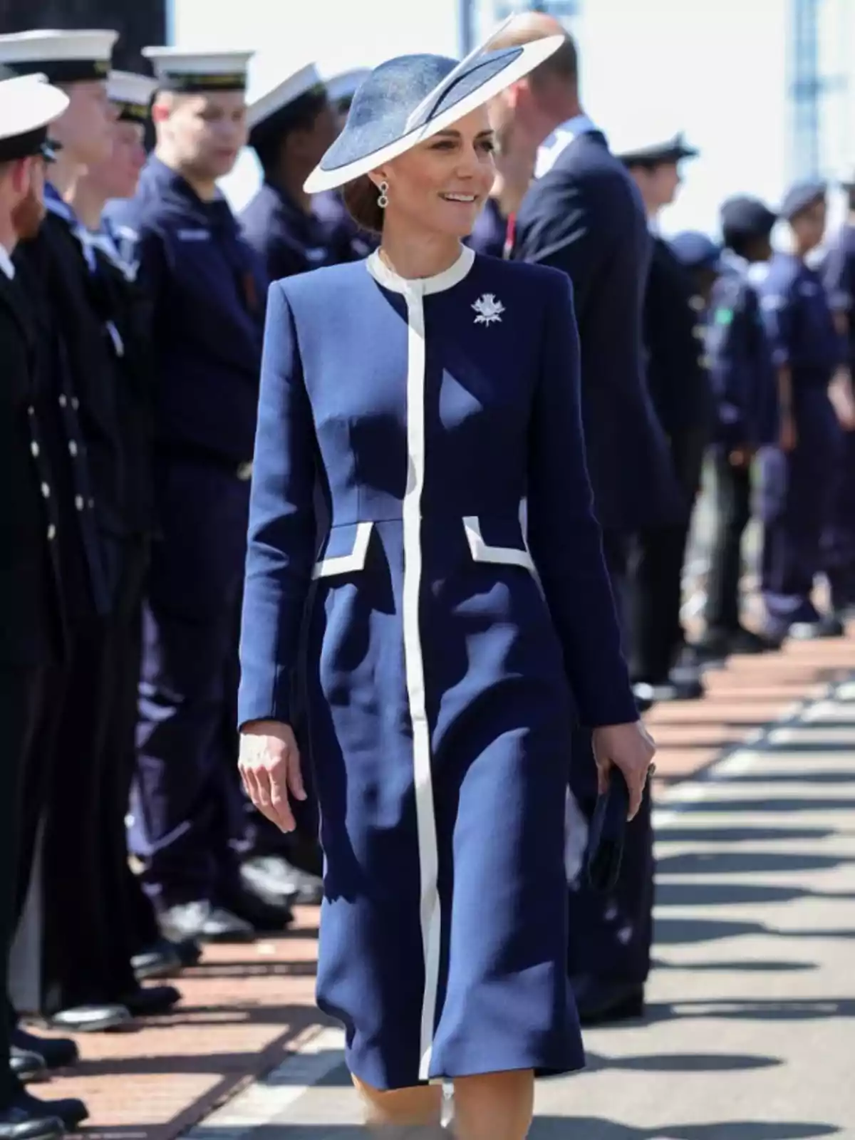 A woman in an elegant blue dress and matching hat walks outdoors while uniformed people are lined up around her. A woman in an elegant blue dress and matching hat walks outdoors while uniformed people are lined up around her.