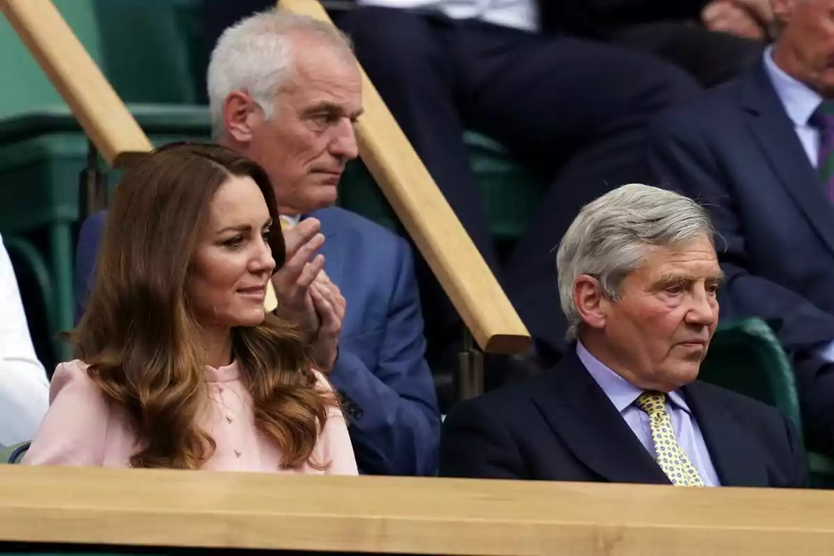 Personas sentadas en una grada, Kate Middleton con cabello largo y suelto y Michael Middleton con cabello canoso, ambos mirando hacia adelante. Personas sentadas en una grada, Kate Middleton con cabello largo y suelto y Michael Middleton con cabello canoso, ambos mirando hacia adelante.