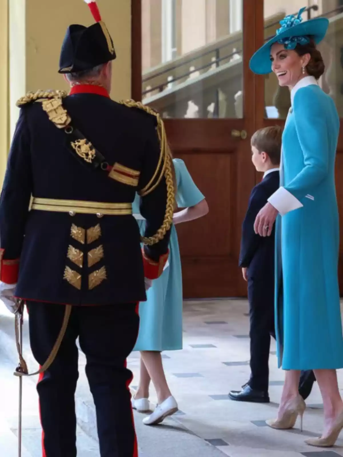 Una dona elegantment vestida de blau amb un barret a joc somriu mentre conversa amb un home uniformat de gala i dos nens en un edifici d’aspecte oficial. Una dona elegantment vestida de blau amb un barret a joc somriu mentre conversa amb un home uniformat de gala i dos nens en un edifici d’aspecte oficial.