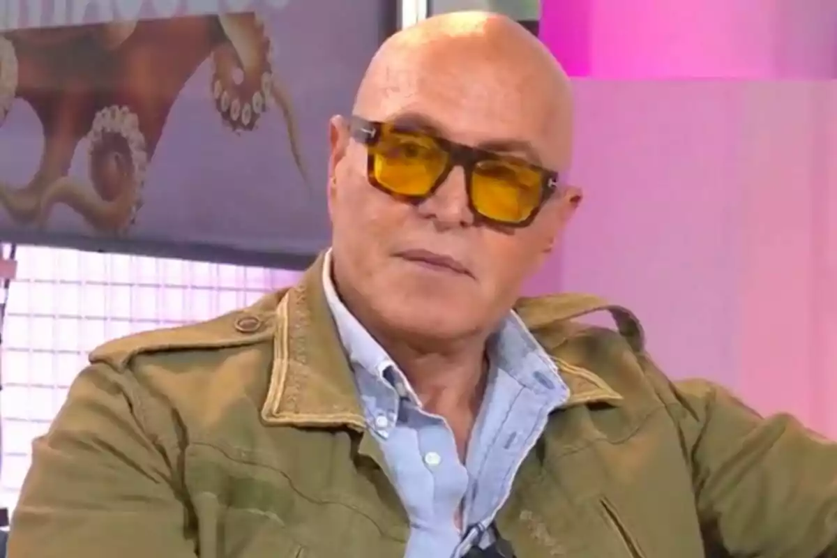 Kiko Matamoros calvo con gafas de sol amarillas y chaqueta verde está sentado en un entorno iluminado con luces rosadas y un cartel de fondo que muestra el programa 'Tentáculos'. Kiko Matamoros calvo con gafas de sol amarillas y chaqueta verde está sentado en un entorno iluminado con luces rosadas y un cartel de fondo que muestra el programa 'Tentáculos'.