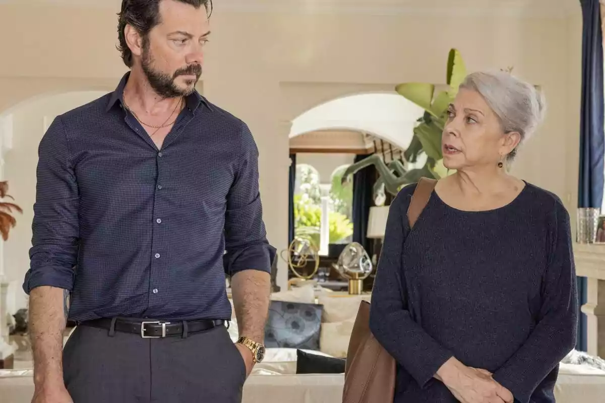 Álvaro Morte de barba y Kiti Mánver de cabello canoso conversan en una sala elegante con plantas y decoración moderna al fondo en Dos Tumbas