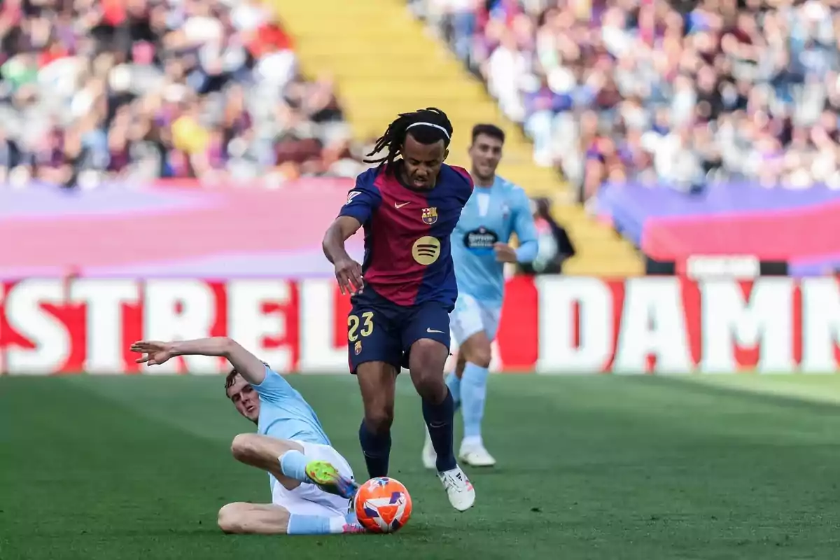 A player wearing number 23 from the team in blue and maroon controls the ball while a player from the opposing team, dressed in light blue, attempts to stop him by sliding on the grass. A player wearing number 23 from the team in blue and maroon controls the ball while a player from the opposing team, dressed in light blue, attempts to stop him by sliding on the grass.