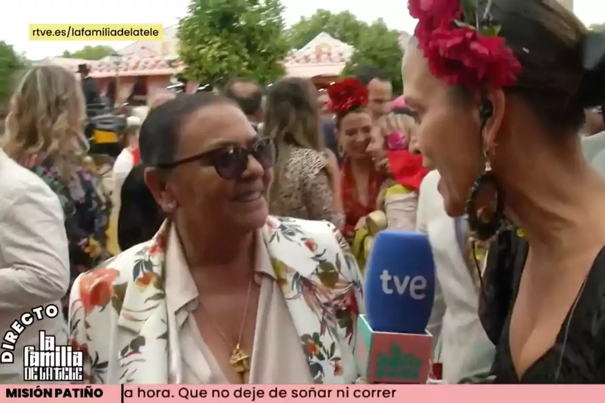 Una reportera entrevista a María del Monte en un esdeveniment a l'aire lliure, envoltades de persones vestides amb vestits acolorits i adorns florals. Captura de 'La Família de la Tele' Una reportera entrevista a María del Monte en un esdeveniment a l'aire lliure, envoltades de persones vestides amb vestits acolorits i adorns florals. Captura de 'La Família de la Tele'