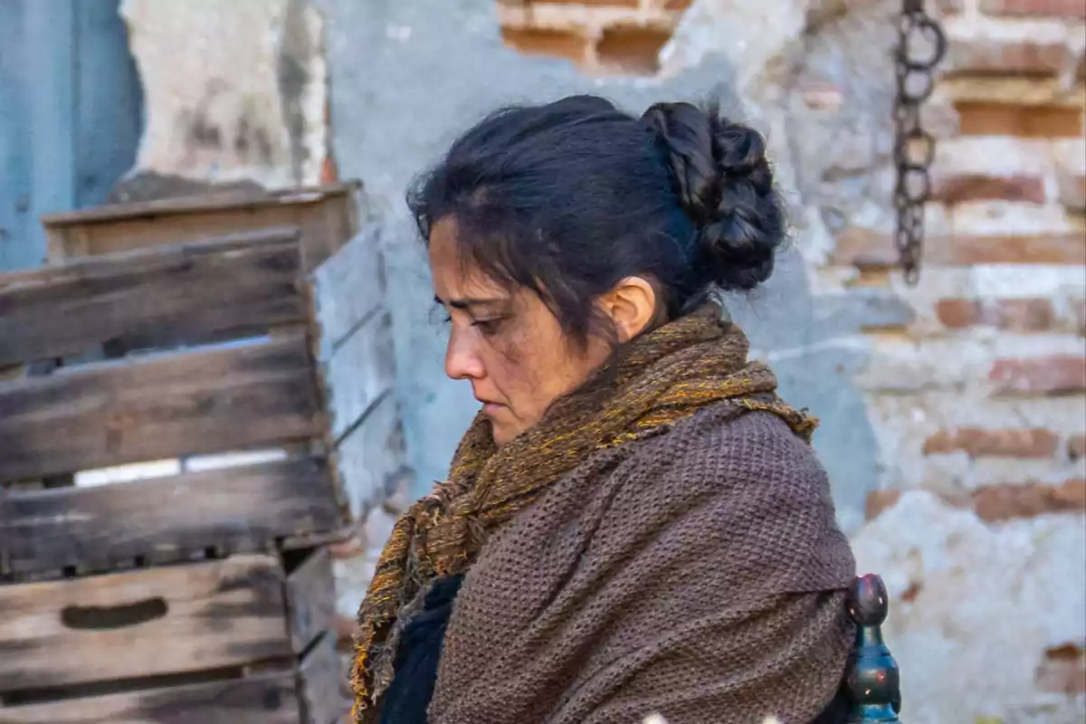 Alícia amb els cabells recollits i bufanda marró, de perfil, davant d'un fons de caixes de fusta i paret de maó. Captura de 'La Promesa'. Alícia amb els cabells recollits i bufanda marró, de perfil, davant d'un fons de caixes de fusta i paret de maó. Captura de 'La Promesa'.