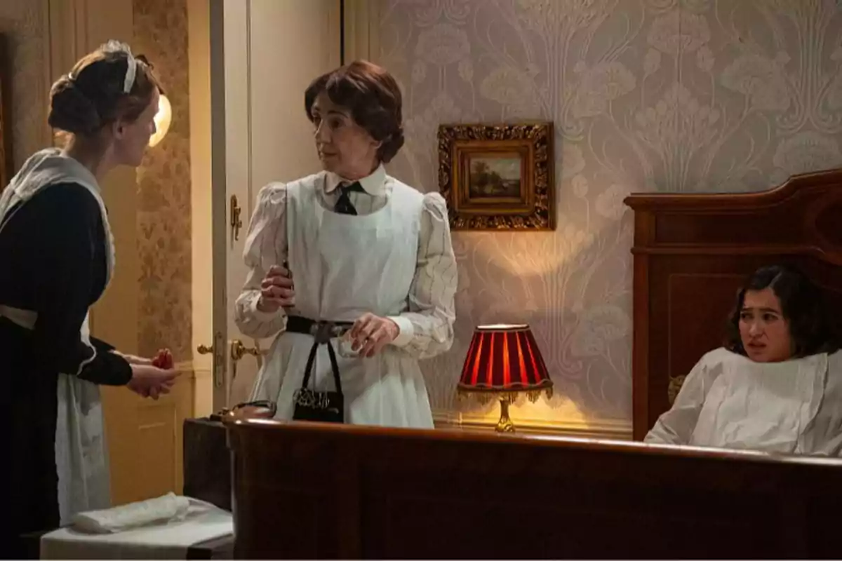 Pía, Emília i Catalina en una habitació antiga, dues d’elles amb uniforme de minyona i una asseguda al llit amb expressió preocupada, al costat d’una làmpada de taula encesa i una paret decorada amb paper pintat. Captura de ‘La Promesa’. Pía, Emília i Catalina en una habitació antiga, dues d’elles amb uniforme de minyona i una asseguda al llit amb expressió preocupada, al costat d’una làmpada de taula encesa i una paret decorada amb paper pintat. Captura de ‘La Promesa’.